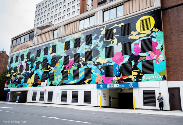 Artist EEPMON stands in front of a building featuring one of his large, colourful murals. EEPMON is standing in the bottom right of the photo. | L'artiste EEPMON se tient devant un bâtiment orné d'une de ses grandes fresques murales colorées. EEPMON se trouve en bas à droite de la photo.