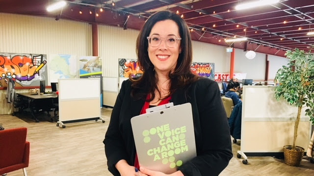 Tanya Woods smiles at the camera while holding a clipboard with the words "One voice can change a room." | Tanya Woods sourit à la caméra tout en tenant un bloc-notes sur lequel on peut lire « Une seule voix peut changer l'ambiance d'une pièce » (mais écrit en anglais).