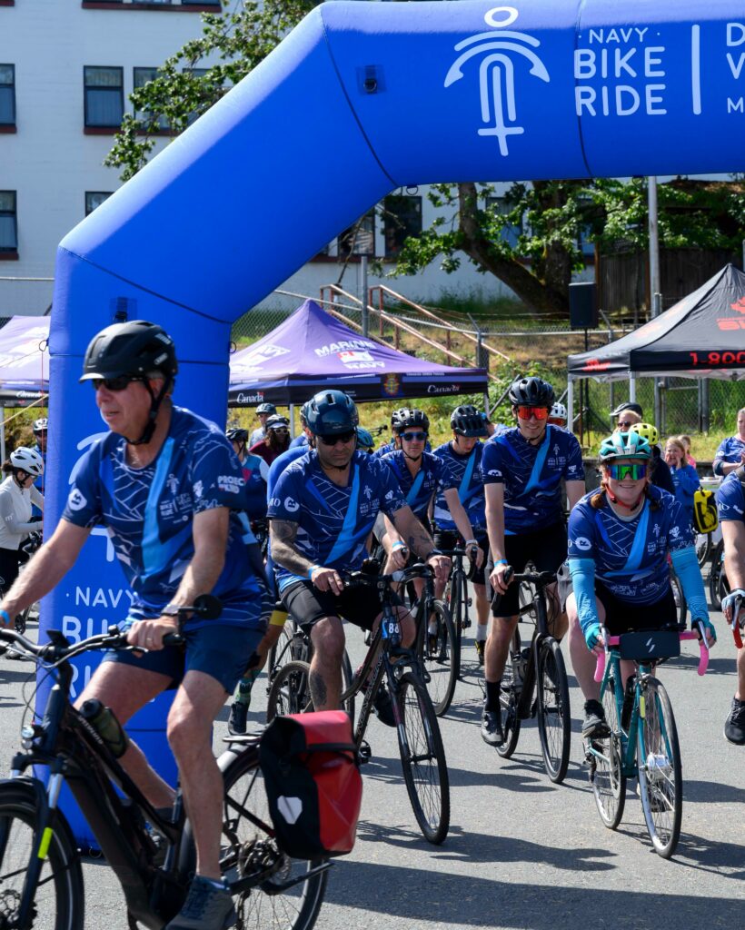 Cyclist wearing Navy Bike Ride shirts depart under a bright blue arch. | Des cyclistes vêtus de t-shirts du Défi-vélo de la Marine s'élancent sous une arche d'un bleu éclatant.