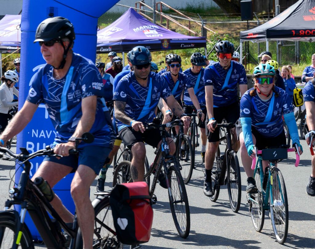 Cyclist wearing Navy Bike Ride shirts depart under a bright blue arch. | Des cyclistes vêtus de t-shirts du Défi-vélo de la Marine s'élancent sous une arche d'un bleu éclatant.